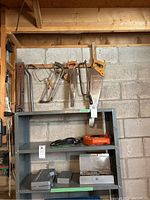Wide view of tools on a gray shelving unit including hacksaws, hand saws, small metal box, case with drill bits, extension cords, and wooden level.