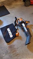 Worx WG502.2 leaf blower and black collection bag laid on a concrete floor, showing side angle of tool and bag attachment ring.