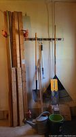 Photo showing assorted wood planks clamped, long-handled yard tools including shovel, brooms, rake, watering can, buckets, and boxed gutter guard arranged against a wall in a garage.