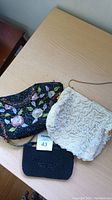 Three theatre clutches on wooden surface including beige crocheted floral clutch, black sequin floral clutch, and small black beaded clutch.