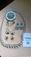 All jewelry pieces and compact mirror displayed on wood surface including clear bead necklace and 5 pairs of earrings