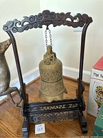 Full view of the brass hanging bell suspended from a carved dark wooden stand with ornamental scrollwork.