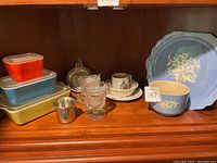 Shows assorted kitchenware including ceramic blue floral patterned bowl and plate, vintage Tupperware containers, stainless steel cup, glass dessert dishes, and Peter Rabbit themed cup and plates.