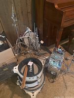 Full view of drum components including stacked white drums, cymbal stands, and drumsticks with tagged label showing Roland branding under drumhead.