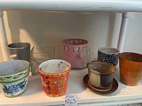 Photo showing assortment of seven decorative flower pots in different styles and materials on a shelf.