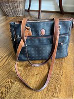 Black handbag with brown leather straps resting on wooden floor, showing front and side pockets and the distinct Kenzia brand pattern on exterior.