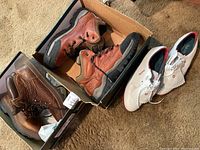 Three pairs of men's shoes on floor: two pairs Timberland boots in brown leather in boxes and one pair white FootJoy cleats outside box.