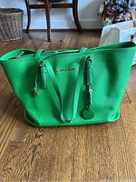 Front view of a bright green Michael Kors handbag on wooden floor, showing gold-tone logo and zipper pull.