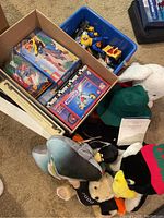 Photo showing blue bin with toy cars and stuffed animals including a white rabbit and black stuffed with red and green clothing. Box with K'NEX kits and LEGO sets visible.