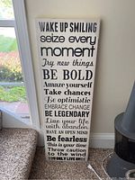 Photo of a tall white rectangular wall art panel with black motivational text placed on the floor against a wall near a window and a chair.