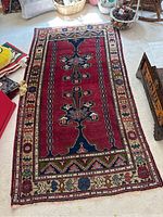 Rectangular woven runner rug on carpeted floor with red primary color and geometric floral patterns in blue and cream, displayed with surrounding home decor items.