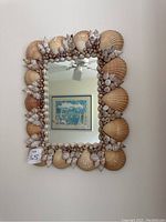 Full view of the rectangular mirror with seashell frame hung on a white wall, reflecting a ceiling fan and a framed picture.