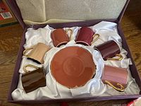 Set of seven ceramic mugs in various earthy colors with gold handles arranged in a box around a ceramic plate.
