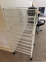 Front angle of white wire shelving unit showing four wire basket shelves, white frame, and black caster wheels.