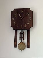 Full view of wooden wall clock with pendulum showing dark wood clock face with gold-tone numerals and two hands.