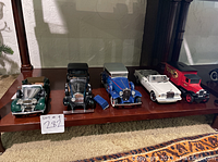 Front view of five Franklin Mint antique toy cars on a wooden shelf including a green vintage car, black sedan, blue convertible coupe, white convertible, and red model truck.
