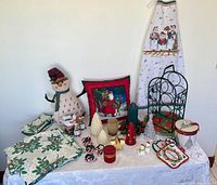 Photo showing assorted holiday-themed items arranged on table including fabric linens with holly design, various Christmas candles, light-up decorations, Christmas-themed pillow and apron, metal wine rack, salt and pepper shakers, gift box, advent snowman calendar, potholders, napkin holders, and kitchenware items.