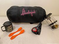 Sleeping bag, travel mug, gas lantern, orange spork utensils, and headlamp arranged on a flat surface for overview.