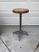 Full view of barstool with wooden round seat and metal base with footrest on concrete floor.