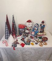 Full overview of Christmas decor items arranged on cloth-covered table including fabric cone trees, Santa holder, nutcracker, assorted ornaments, mugs, snowman figurine, and decorative tins.
