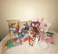 Photo showing Easter eggs, decorative pillow with bunny silhouette and stain, Easter bags, bunny ears, plush animals, and other seasonal items arranged on a table