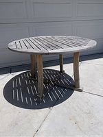Round weathered outdoor wooden patio table shown from side angle on concrete surface.