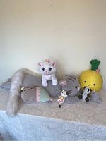 Full lot front view showing all plush toys arranged on a table with light lace cover, including large Pusheen cat, white cat, pineapple, panda, and smaller animal plushies.