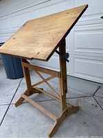 Angled view of a vintage wooden drafting table with visible wear and scratches on the surface and frame.