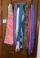 Eight scarves hanging on rack, showing variety of colors and patterns including purple, pink print, teal metallic, black and white snake print, solid purple, and more.