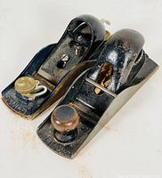 Top side view of two antique Stanley block planes showing brass knobs and wooden handles with japanning finish, placed on white surface
