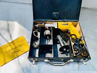 Overview of the Dewalt corded drill in its open metal carrying case with several accessories including a manual, chuck attachments, and auxiliary handle.