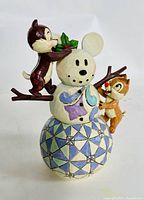 Front view showing Chip placing holly on Mickey Mouse snowman while Dale supports from the side branch; colorful painted geometric base.