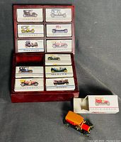 Top-down view of 11 diecast mini antique car boxes arranged in a wooden holder, with one red and orange miniature car outside its box.