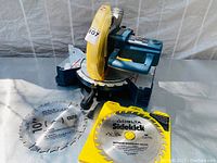 Photo showing Ryobi miter saw with yellow blade guard, two circular saw blades laid out, one Ryobi and one Delta Sidekick with visible details.