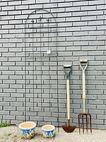 Photo showing two ceramic planters, black steel garden trellis, garden hoe, and pitchfork against a gray brick wall.