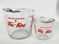 Two vintage Anchor Hocking Fire King glass measuring cups, one larger with a handle and one smaller without a handle.