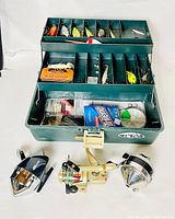 Photo of the metal green tackle box open showing sections filled with assorted colorful fishing lures and accessories, placed behind three vintage fishing reels. The reels include two black and chrome conical shaped reels and one spinning reel with cream and gold color.