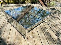 Square vintage smoked glass top coffee table with chrome frame on outdoor wooden deck with reflections of trees.
