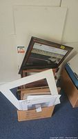 Two cardboard boxes holding assorted wooden picture frames of various sizes, some matting, and foam boards; frames are mostly rectangular with wood finishes and white matting present