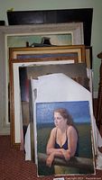 Stack of framed and unframed paintings showing female portrait in dark top leaning on a railing