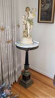 Photo showing full pedestal table with white marble top and dark ornate metal base supporting it. Statue on top is not included.