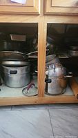 Photo of wooden cabinet lower section storing multiple stainless steel pots with lids, frying pans, colander, and baking pans.
