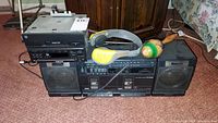 Wide shot showing all items together: Sony boom box with detachable speakers, stacked Acura CD car radio and another audio unit, Sony AM/FM Sports Walkman headphones, and a wooden percussion mallet.
