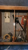 Photo of garden tools including hedge trimmer, extension cord, hand tools, and other accessories on a wooden rack against a wall