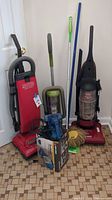 Three upright vacuums, handheld vacuum, Quickie mop and dust mop seen together against wall on patterned floor.