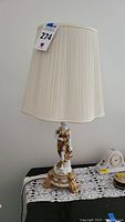 Full view of the vintage porcelain Roman figural woman lamp with cream pleated shade on a table with lace doily and other small household items.