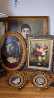 Photo showing four framed artworks: oval painting of Jesus, rectangular portrait of young man, floral still life painting, and two small oval bird prints in ornate frames.