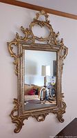 Front view of vintage gilded gold ornate mirror showing full frame, decorative crest at top, and mirror surface reflecting interior room.