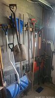 Wide view of yard tools including snow shovels, rakes, broom, dust pan, and shovels leaning against wall in garage.
