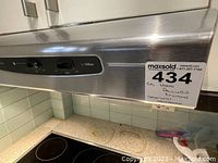 Front and side view of the stainless steel range hood installed under cabinet next to ceramic tile backsplash and windows, with control panel visible showing Allure branding with NuTone.